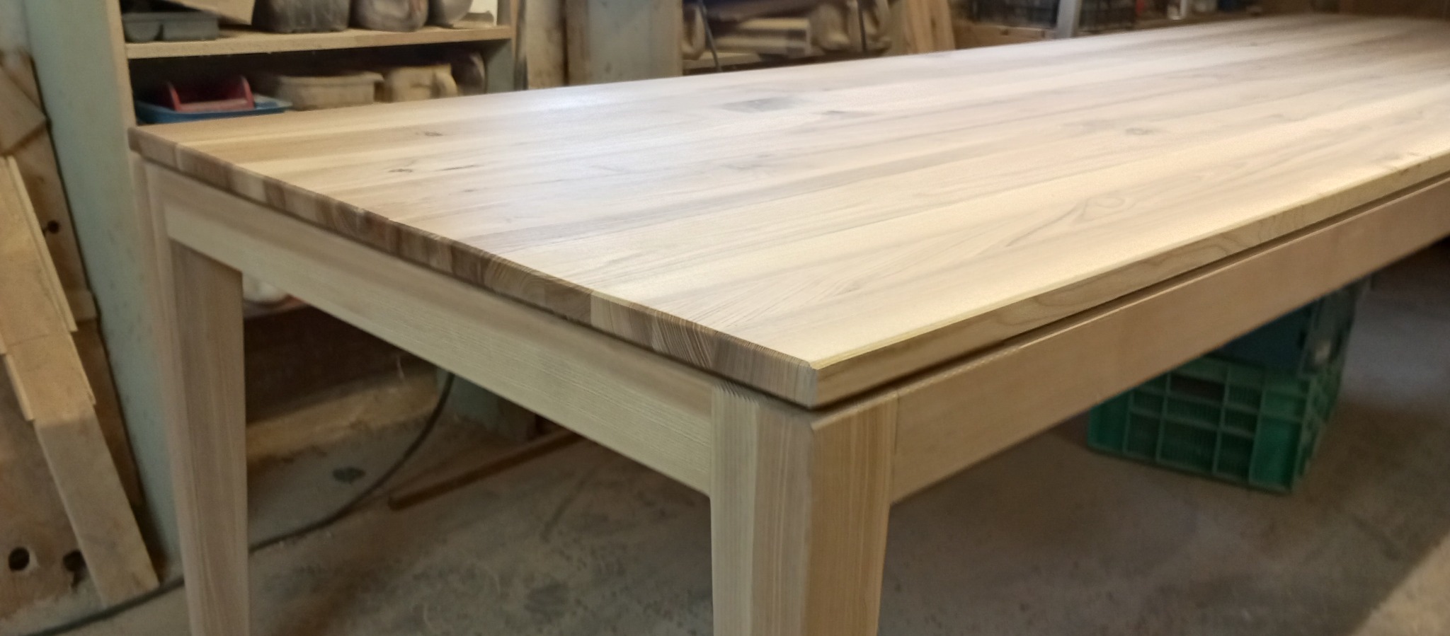 Surowy, drewniany stół z blatem z jasnego drewna, stojący w warsztacie, widoczny pod kątem, z zieloną skrzynką pod spodem i narzędziami na półce w tle.