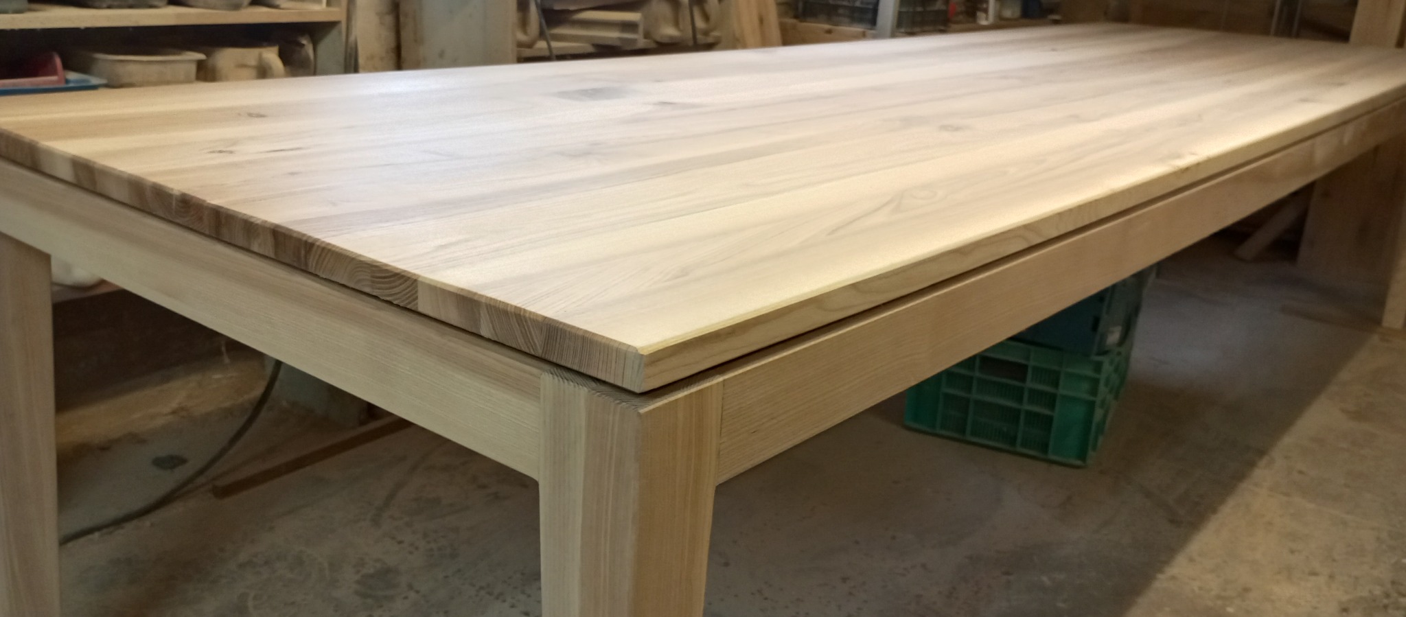 Surowy drewniany stół z blatem z jasnego drewna, widoczne połączenia elementów, zielona skrzynka w tle, warsztat stolarski.