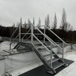 STAL-SPAW Karol Chwajoł - Metalowe schody z platformą i poręczami na dachu budynku, widok na drzewa w tle przy pochmurnej pogodzie.