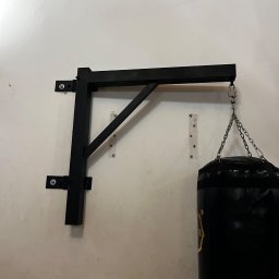 STAL-SPAW Karol Chwajoł - Czarny, stalowy uchwyt ścienny do worka bokserskiego, przymocowany do białej ściany za pomocą śrub; widoczny łańcuch i górna część czarnego worka bokserskiego.