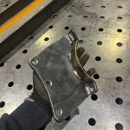 STAL-SPAW Karol Chwajoł - Metalowy element konstrukcyjny z widocznymi spawami, trzymany w rękawicy ochronnej na tle perforowanego stołu spawalniczego.