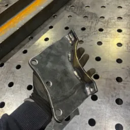 Metalowy element konstrukcyjny z widocznymi spawami, trzymany w rękawicy ochronnej na tle perforowanego stołu spawalniczego.