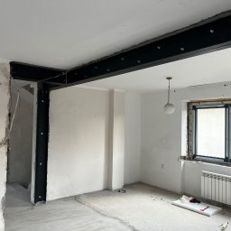 STAL-SPAW Karol Chwajoł - Montaż stalowej belki nośnej w trakcie remontu mieszkania, widoczne elementy mocujące, surowy beton, instalacja elektryczna i okno z grzejnikiem.