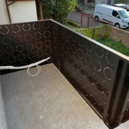 Nowoczesna, ażurowa balustrada balkonowa w kolorze ciemnego brązu z geometrycznym wzorem. Widok z góry, betonowa podłoga balkonu, fragment instalacji elektrycznej.