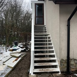 STAL-SPAW Karol Chwajoł - Zewnętrzne, metalowe schody z podestem prowadzące do drzwi wejściowych budynku. Widoczne elementy konstrukcyjne i otoczenie w zimowej scenerii.
