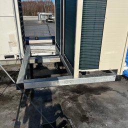 STAL-SPAW Karol Chwajoł - Montaż klimatyzacji na dachu budynku. Widoczna stalowa konstrukcja wsporcza pod agregat, elementy instalacji i fragment dachu. Słoneczny dzień.