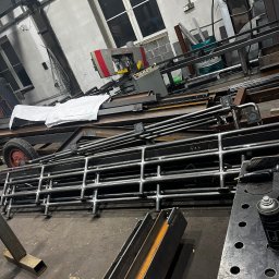 STAL-SPAW Karol Chwajoł - Stalowe konstrukcje w warsztacie: rzędy spawanych, chromowanych rur, profile, narzędzia i maszyna do cięcia metalu w tle. Widoczne plany techniczne.