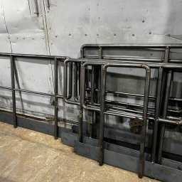STAL-SPAW Karol Chwajoł - Surowe, metalowe balustrady z rur i płaskowników, przygotowane do montażu, opatrzone numerami, ustawione w warsztacie na tle blaszanej ściany.