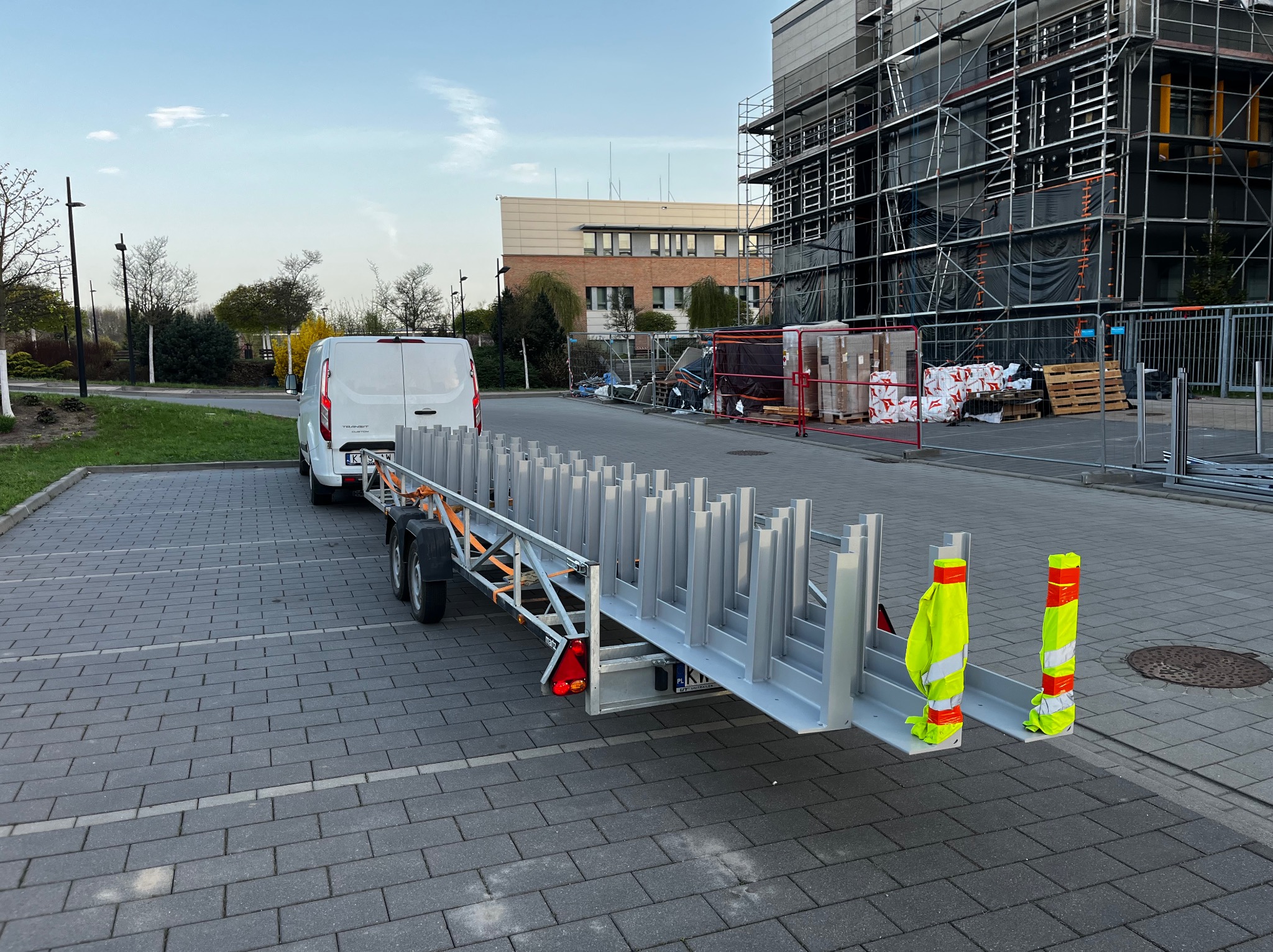 Przyczepa z elementami konstrukcji stalowej, prawdopodobnie do budowy hali, zaparkowana obok budynku w rusztowaniach. Biały bus obok.
