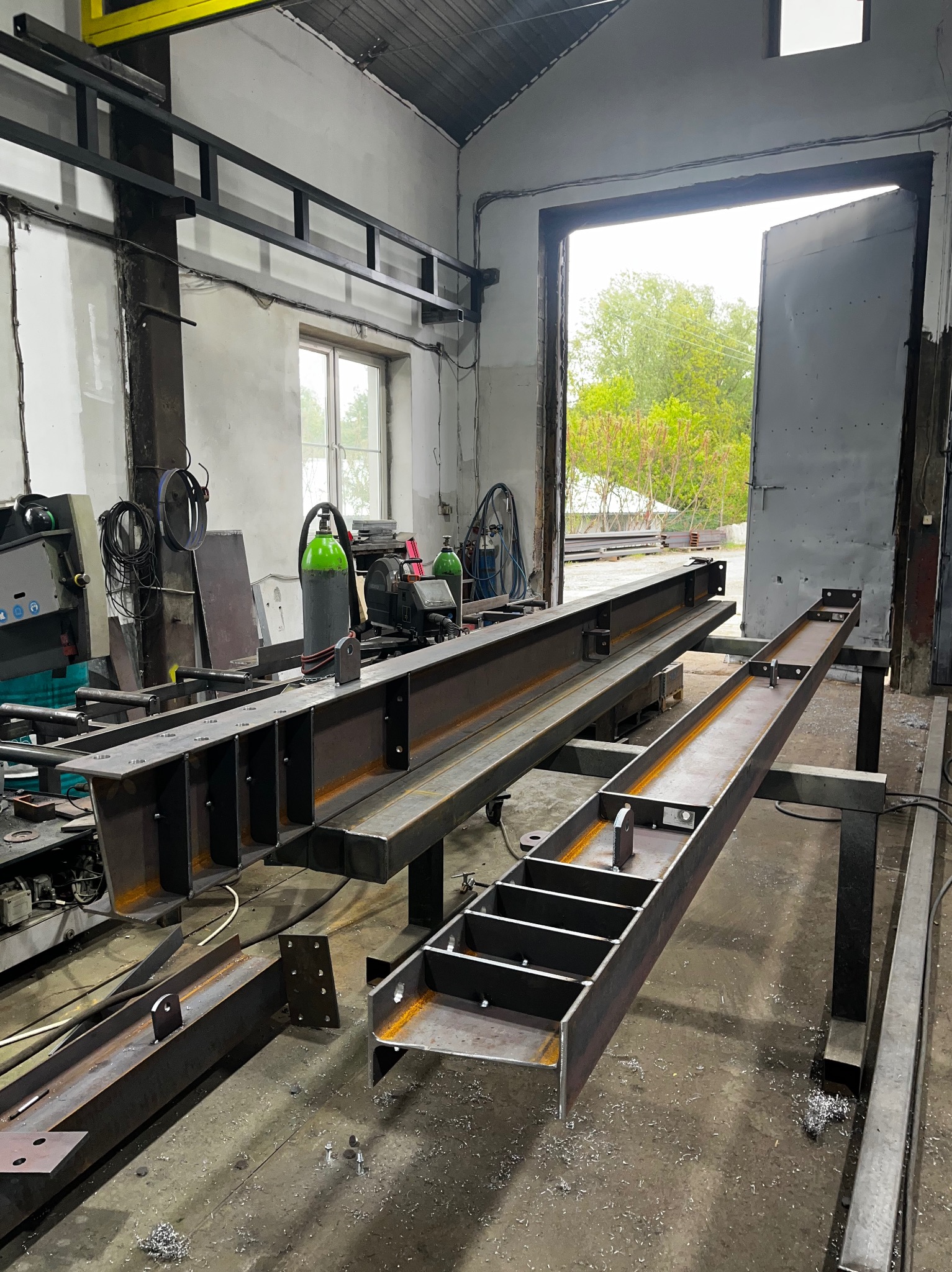 Metalowe konstrukcje hal w warsztacie: spawane elementy, butle z gazem, otwarte drzwi z widokiem na zieleń. Widoczne ślady obróbki metalu.