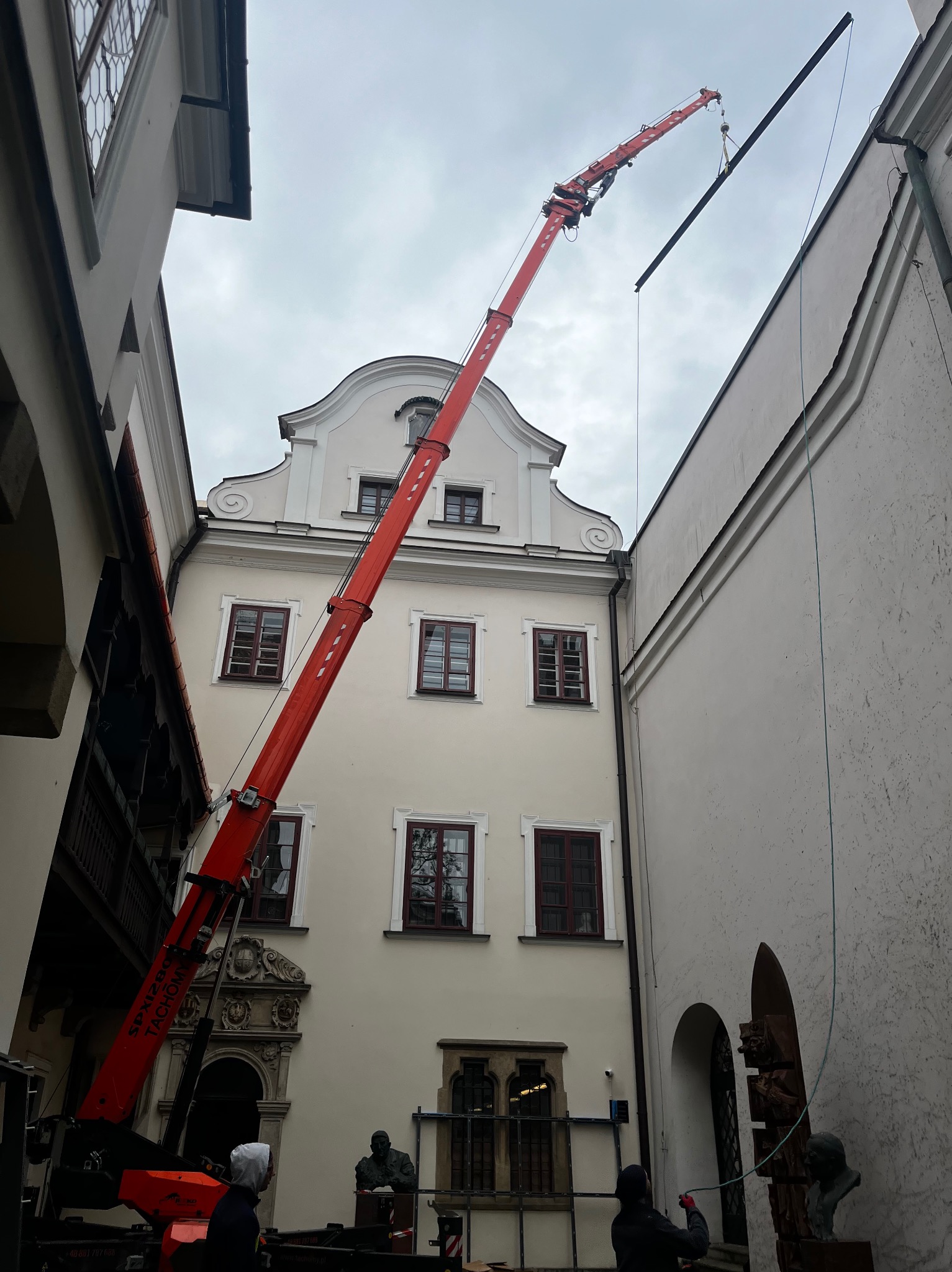 Montaż elementu konstrukcyjnego dźwigiem w wąskiej przestrzeni pomiędzy budynkami, Gdów. Widoczny fragment fasady budynku. Czerwony dźwig w akcji.