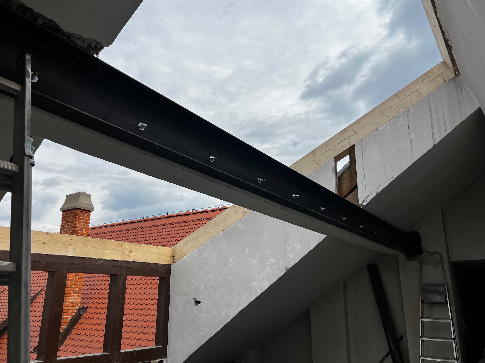 Montaż stalowej belki nośnej w budynku z elementami muru i dachu z cegły. Widoczne śruby mocujące i drabina. Prace ślusarskie w trakcie budowy.