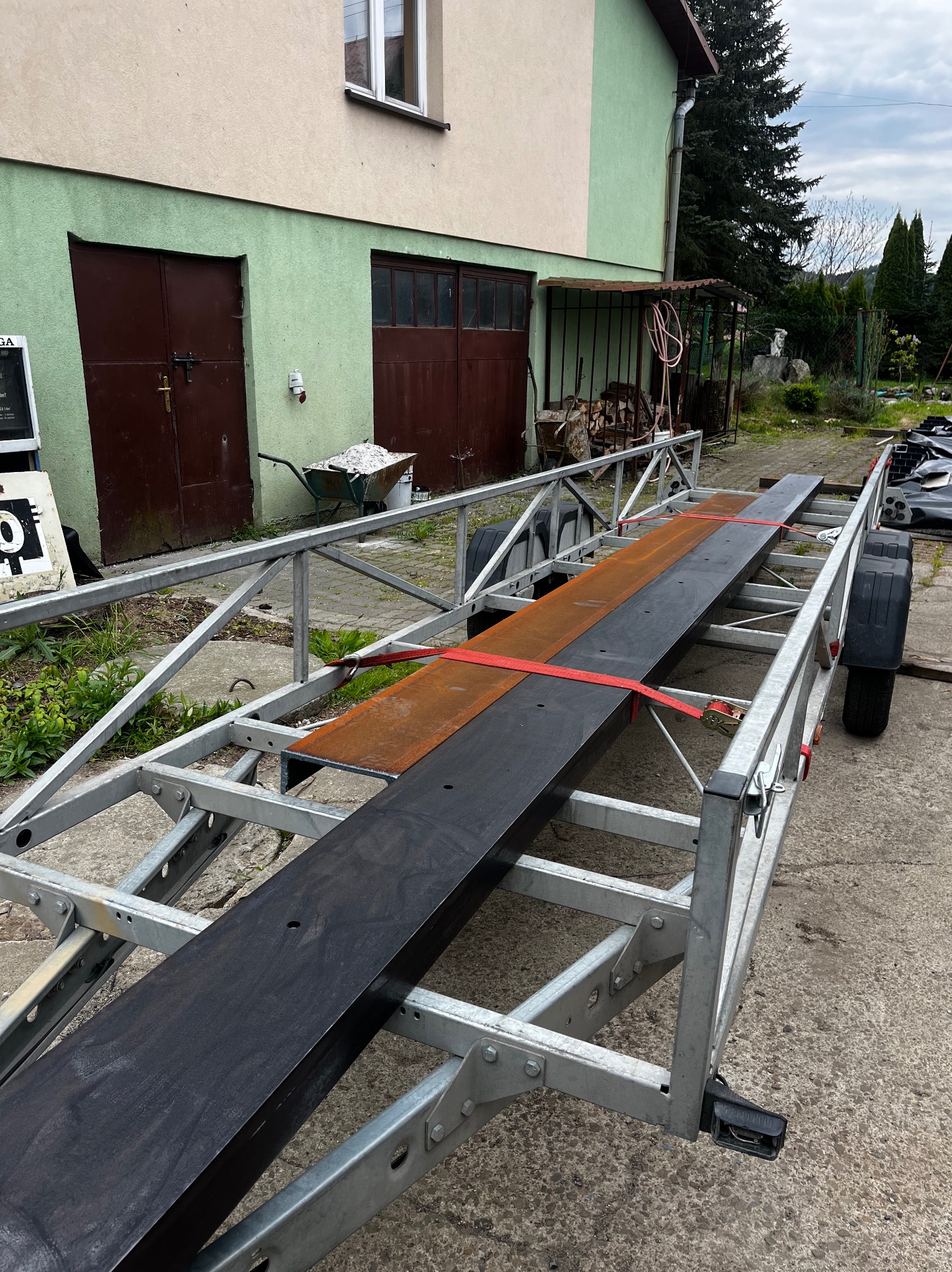 Srebrna, stalowa konstrukcja przyczepy z zamocowanymi elementami metalowymi, zabezpieczonymi czerwoną taśmą. W tle budynek gospodarczy i fragment ogrodu.
