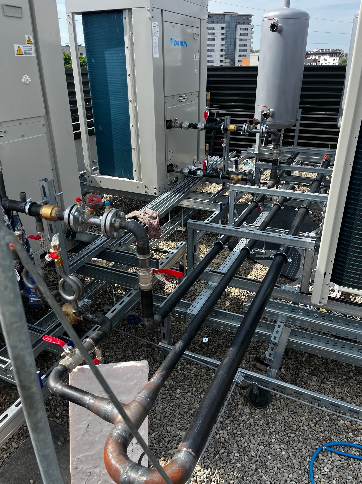 Instalacja przemysłowa na dachu z rurami, zaworami, zbiornikiem i klimatyzatorami Daikin. Widoczne spawane elementy konstrukcji i żwirowe podłoże dachu.