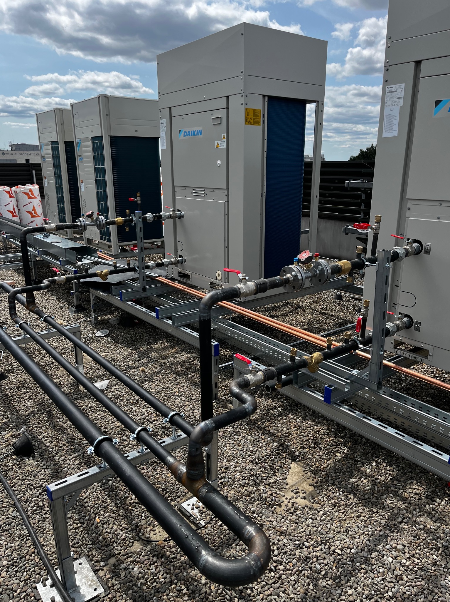 Instalacja przemysłowa: agregaty Daikin połączone rurociągami miedzianymi i stalowymi na dachu pokrytym żwirem, widoczne elementy konstrukcji wsporczej.