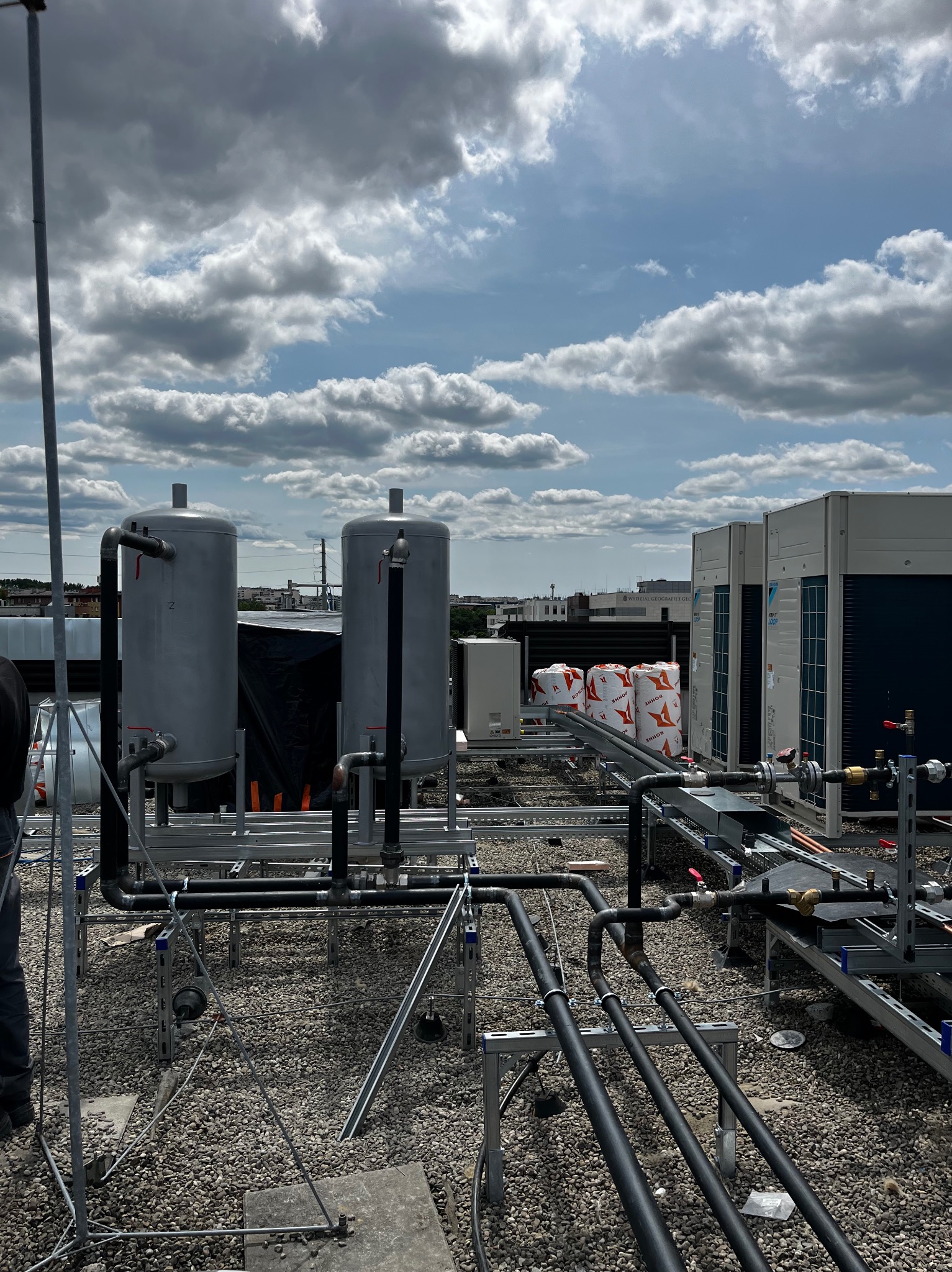 Instalacja przemysłowa na dachu z zbiornikami, rurami i klimatyzatorami. Widoczne elementy konstrukcyjne. Tło stanowi pochmurne niebo i zabudowa miejska.