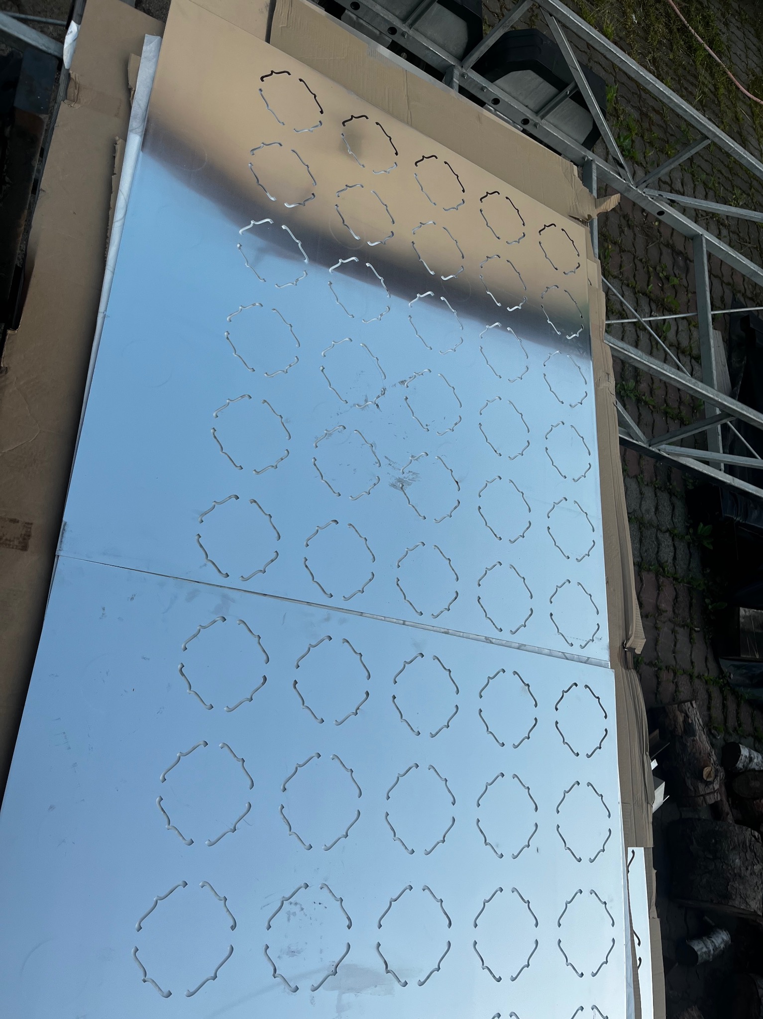 Błyszczące, metalowe arkusze z wyciętymi otworami ułożone na kartonie, obok konstrukcji stalowej. Widoczne nieregularne odbicia światła na powierzchni.