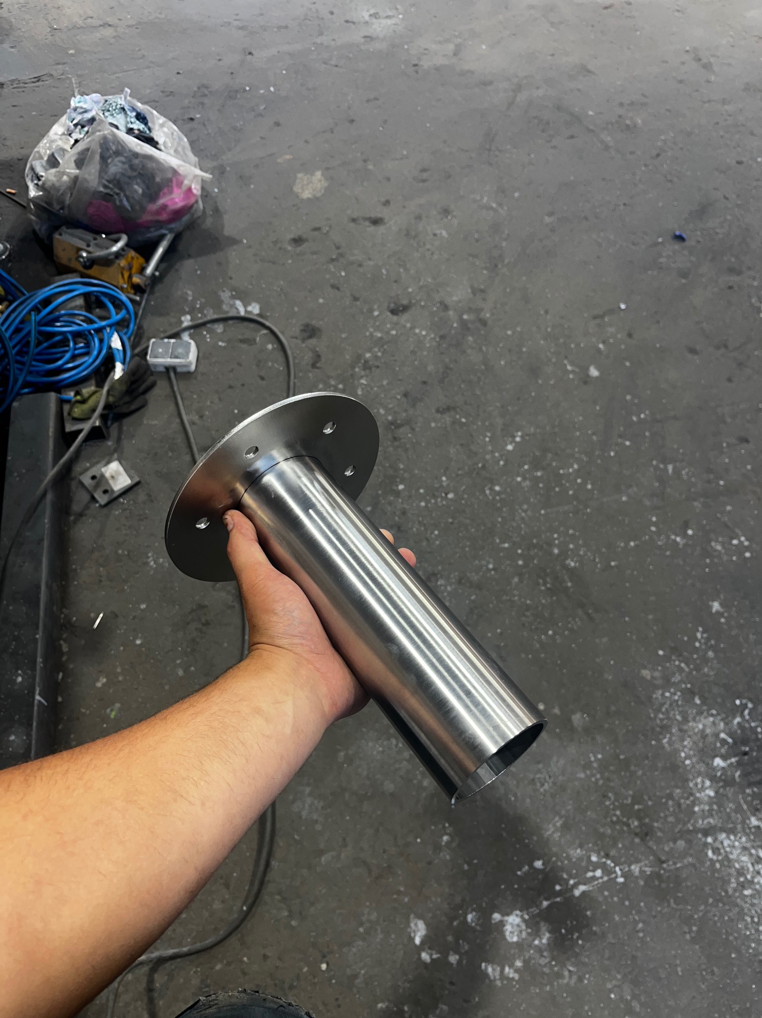 Ręka trzyma gładki, metalowy cylinder z kołnierzem. Element metalowy z otworami na śruby. W tle warsztat z narzędziami i nieporządkiem.