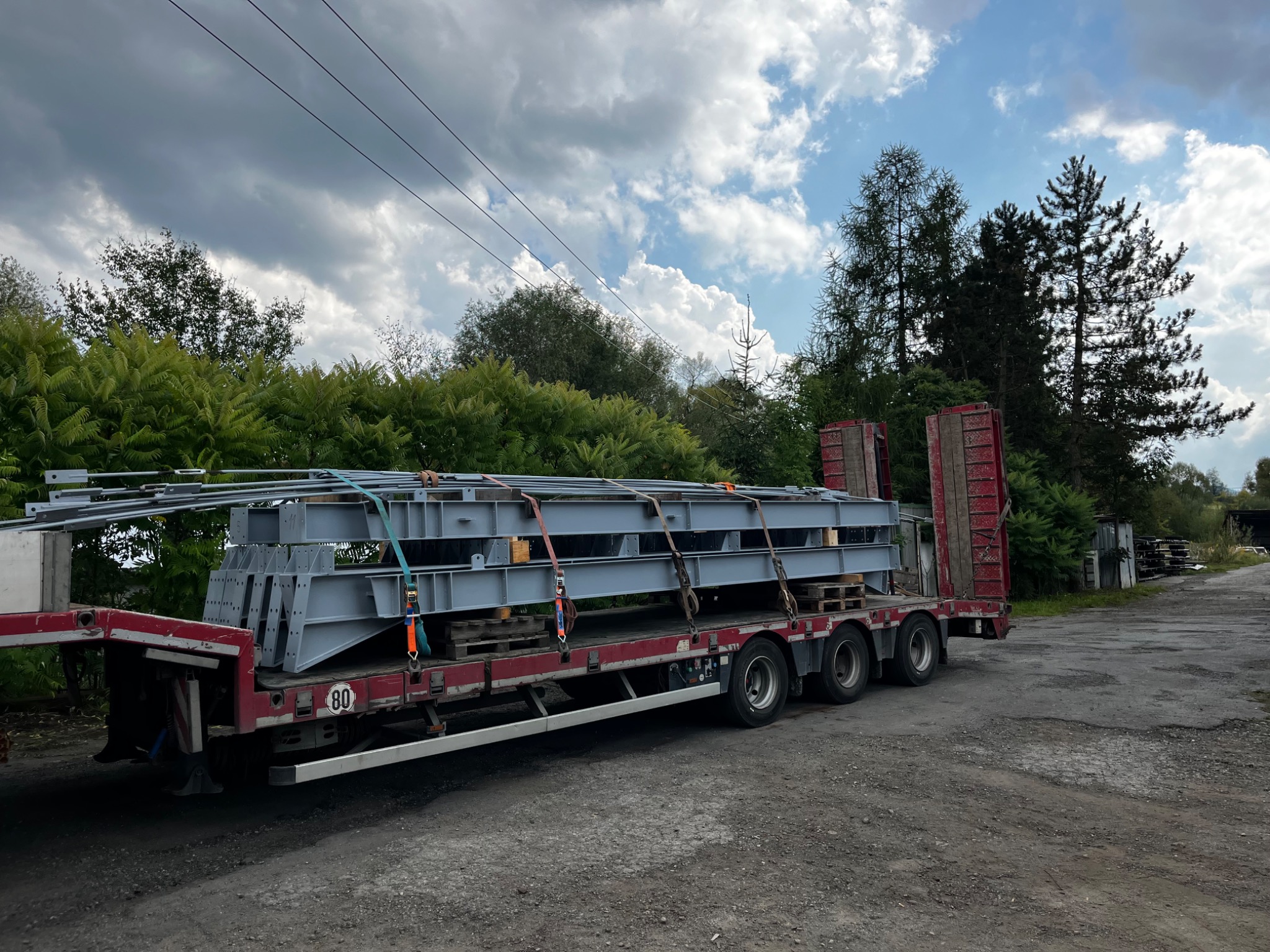 Ciężarówka z naczepą przewozi stalowe elementy konstrukcyjne na paletach, zabezpieczone pasami. Widoczne drzewa i pochmurne niebo w tle.