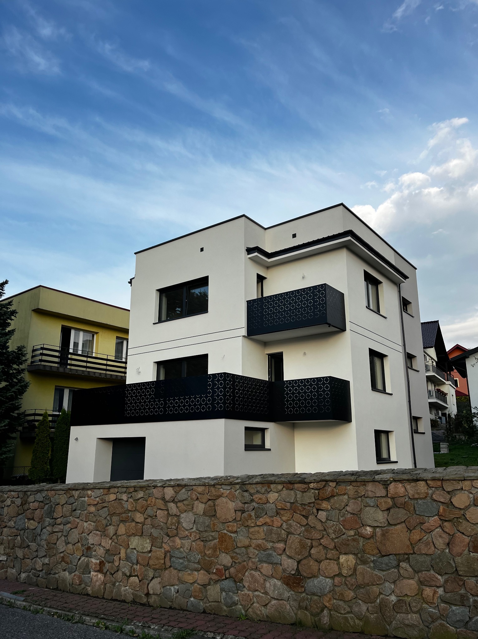Nowoczesny, trzypiętrowy budynek z białymi ścianami i czarnymi, ażurowymi balkonami, widziany zza kamiennego muru. Błękitne niebo z chmurami w tle.