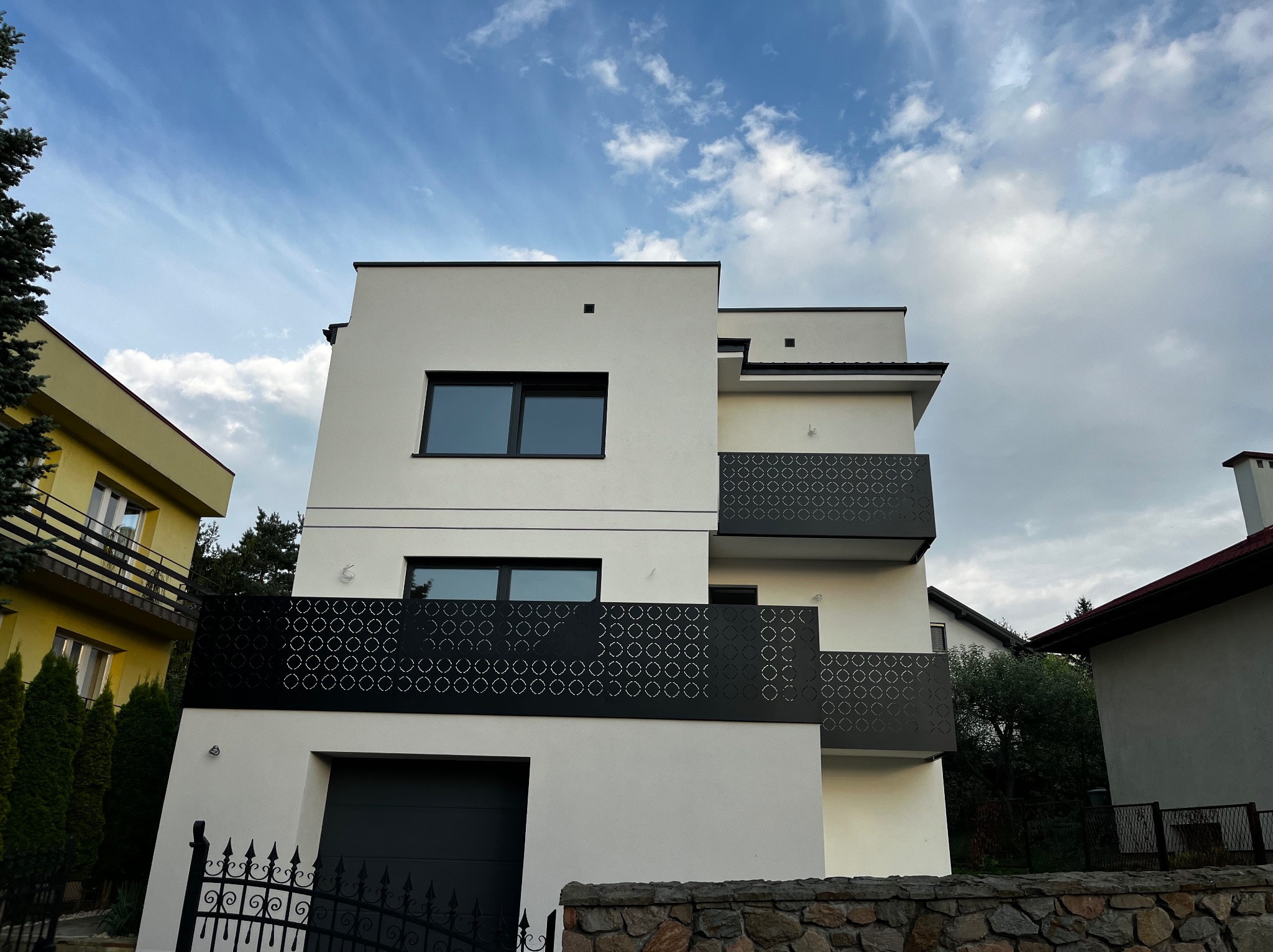 Nowoczesny, trzypiętrowy budynek z białymi ścianami, czarnymi oknami i ażurowymi balustradami balkonów. Widok z dołu na tle błękitnego nieba.
