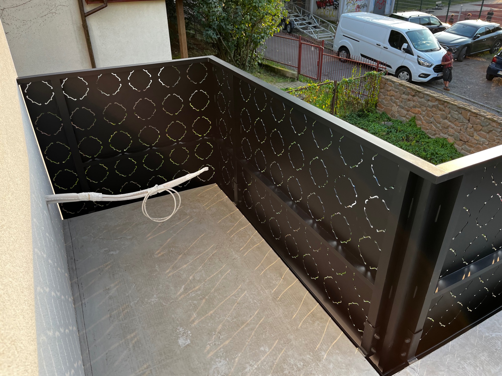 Nowoczesna, ażurowa balustrada balkonowa w kolorze ciemnego brązu z geometrycznym wzorem. Widok z góry, betonowa podłoga balkonu, fragment instalacji elektrycznej.