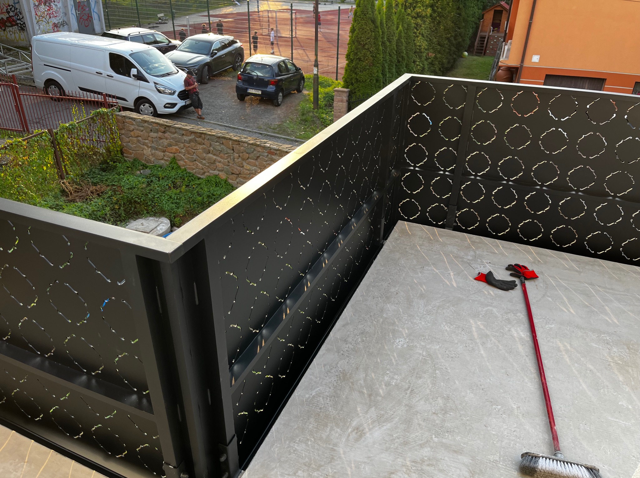 Nowoczesna, czarna balustrada balkonowa z ażurowym wzorem na tle podwórka i zaparkowanych aut. Na balkonie leżą rękawice i szczotka.