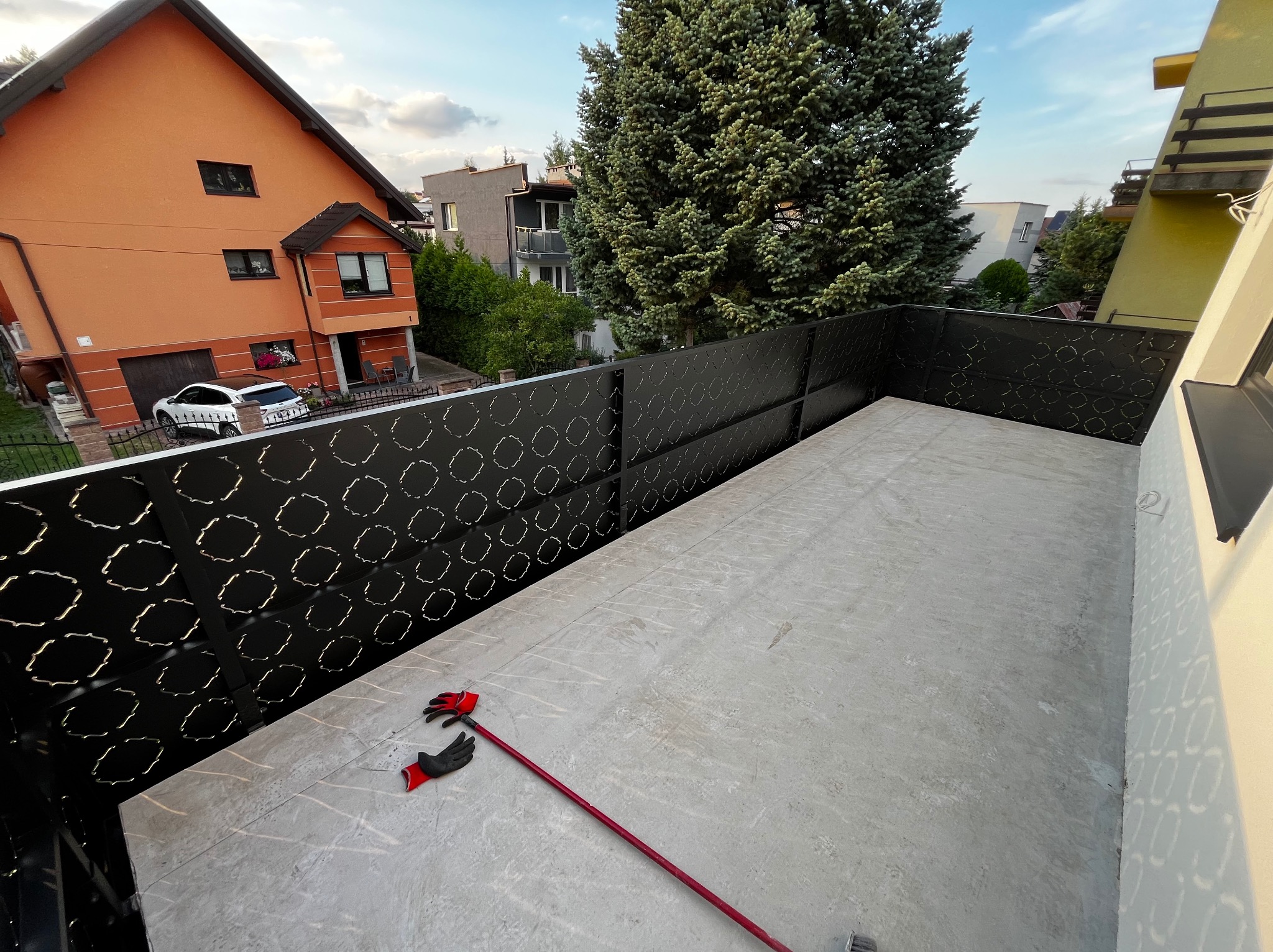 Nowoczesna, czarna balustrada balkonowa z ażurowym wzorem, widok z góry. Na podłodze leżą rękawice robocze i narzędzie. W tle zabudowa miejska.