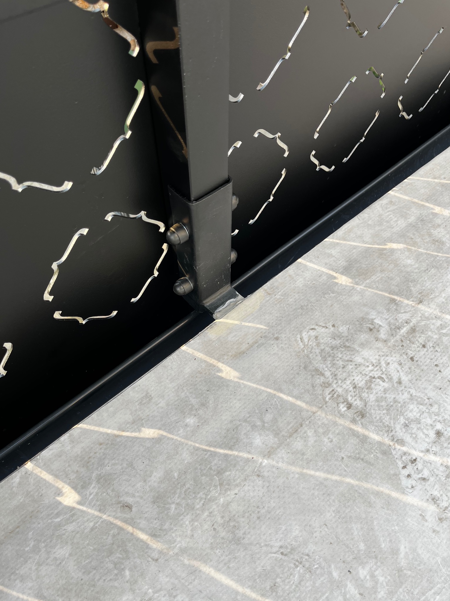 Czarny, metalowy panel z wycinanymi wzorami, przymocowany śrubami do metalowego słupka, osadzony na betonowej posadzce. Widoczne spawanie.