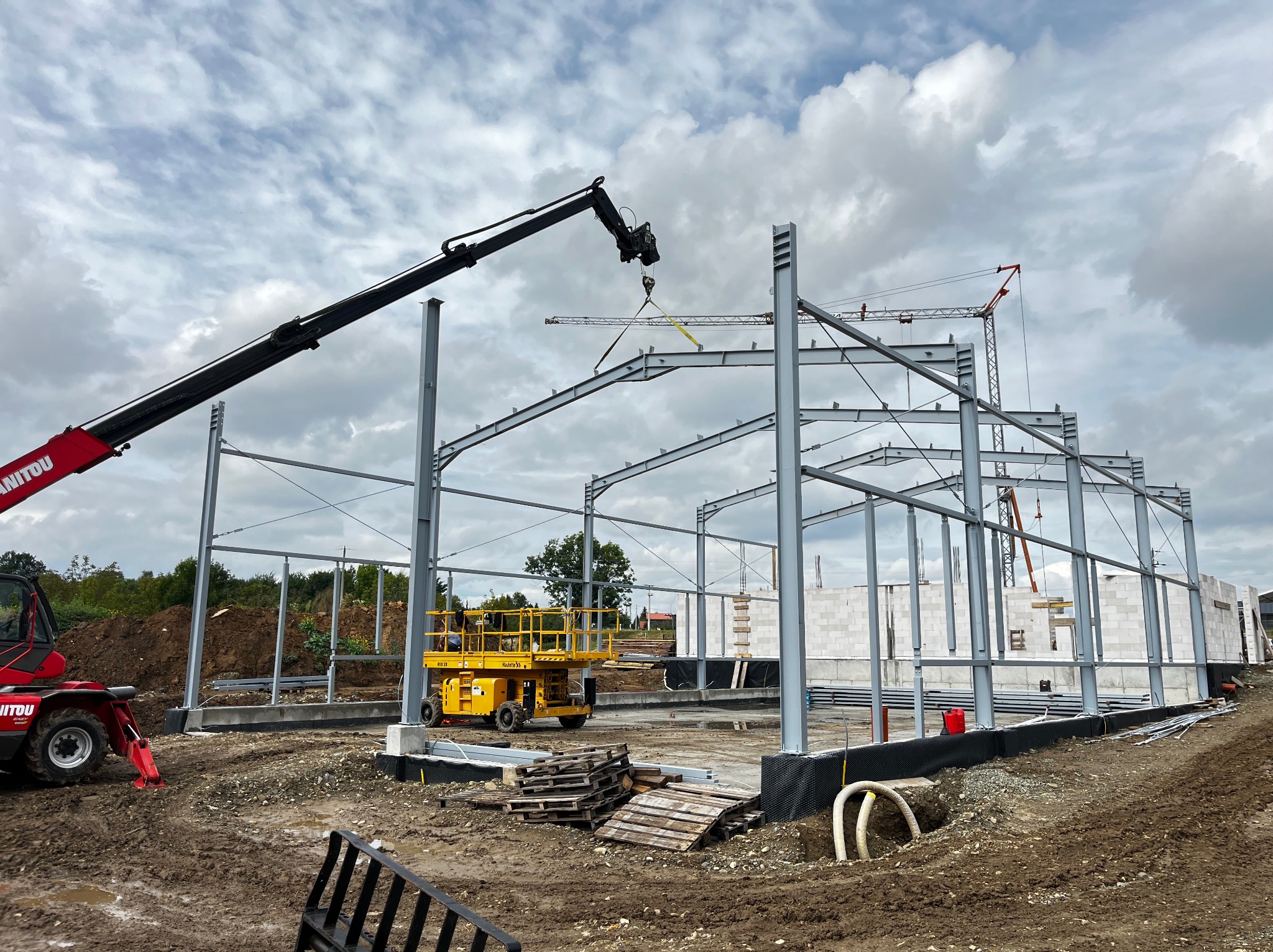 Montaż konstrukcji stalowej hali z użyciem dźwigu teleskopowego Manitou, widoczna żółta platforma robocza i fragment muru z bloczków betonowych.