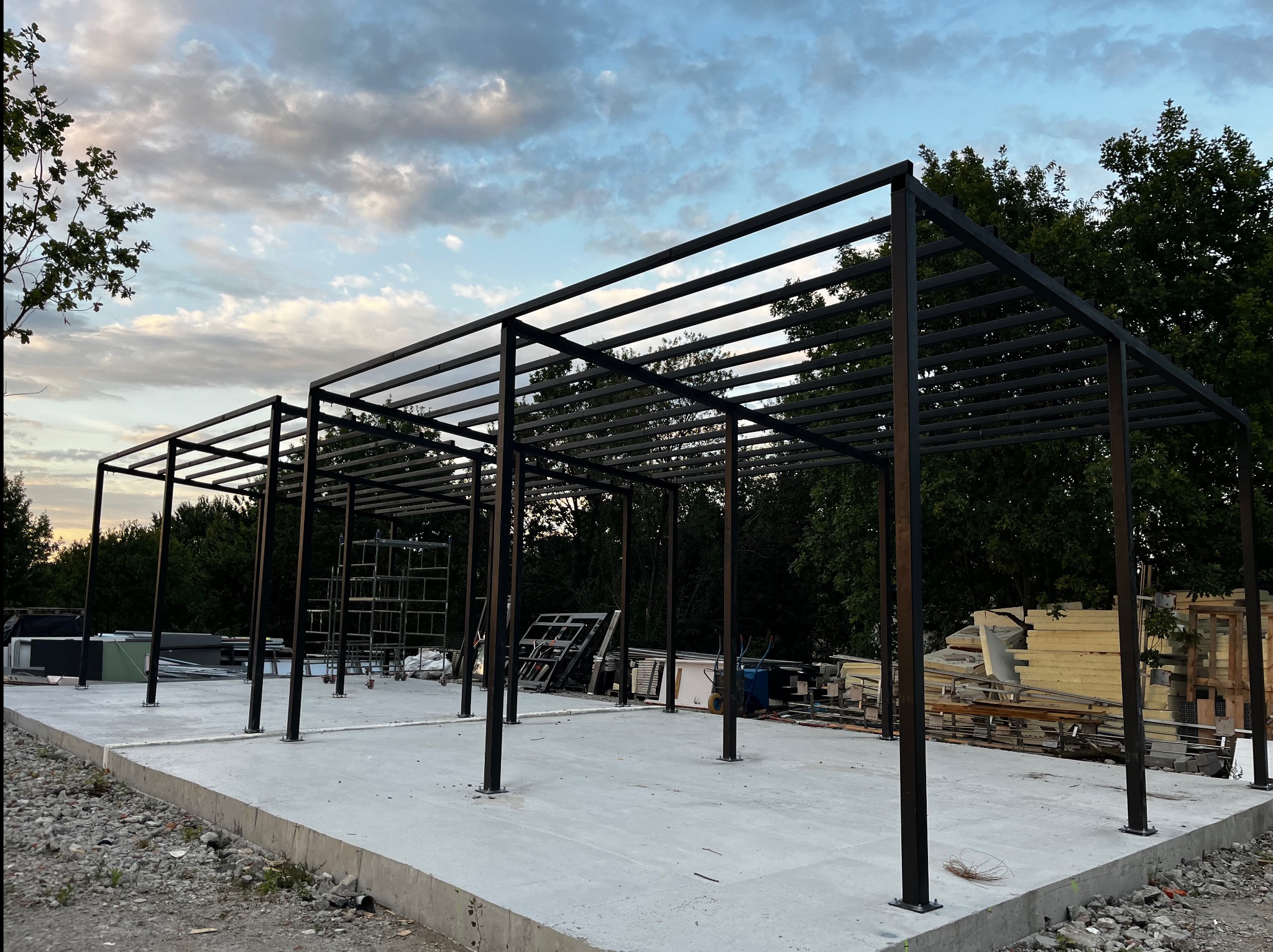 Stalowa konstrukcja hali na betonowej płycie fundamentowej, widoczne rusztowania i materiały budowlane w tle. Błękitne niebo z chmurami w górnej części kadru.