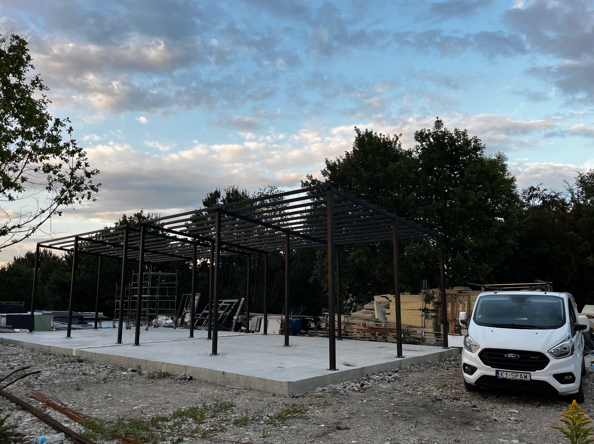 Konstrukcja stalowa z czarnych profili na betonowej podstawie, częściowo zadaszona, z białym busem Ford Transit w tle i zielenią drzew. Widok z zewnątrz.