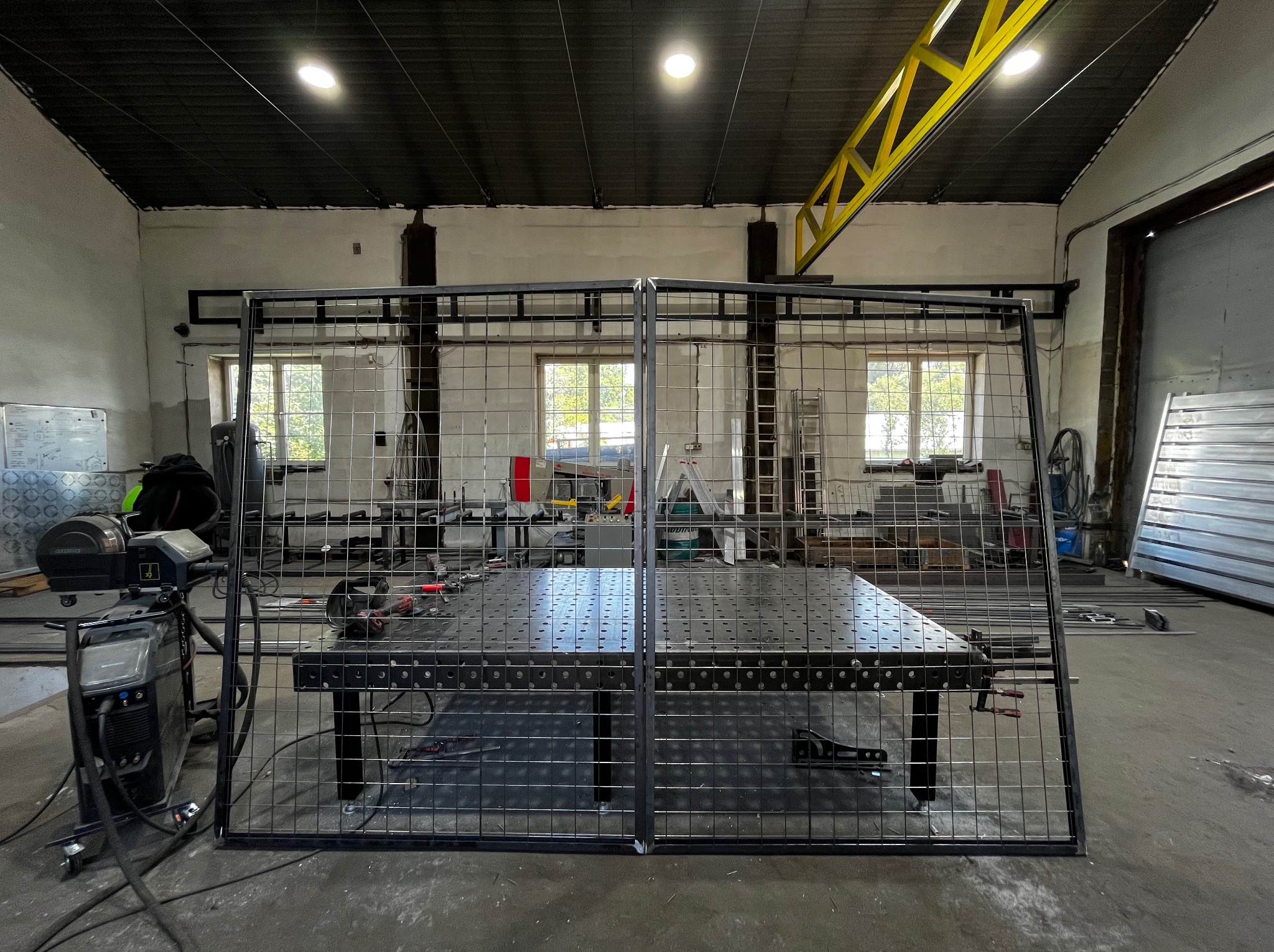 Metalowa konstrukcja kratowa z widocznym stołem warsztatowym w tle, oświetlona światłem dziennym w hali produkcyjnej.