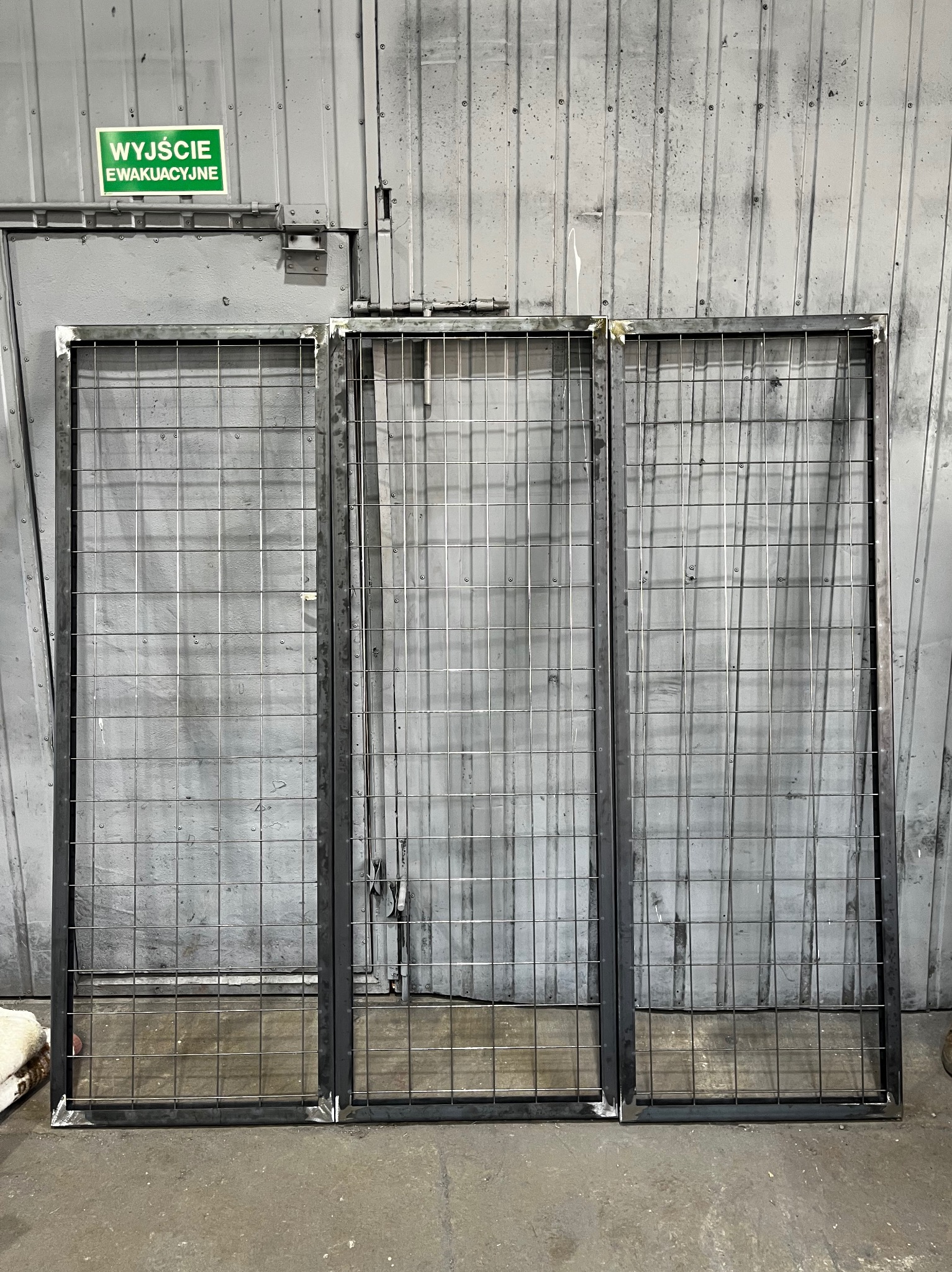 Trzy metalowe ramy z siatką na tle szarej ściany z napisem 'Wyjście Ewakuacyjne'. Konstrukcje spawane, gotowe do dalszego montażu.