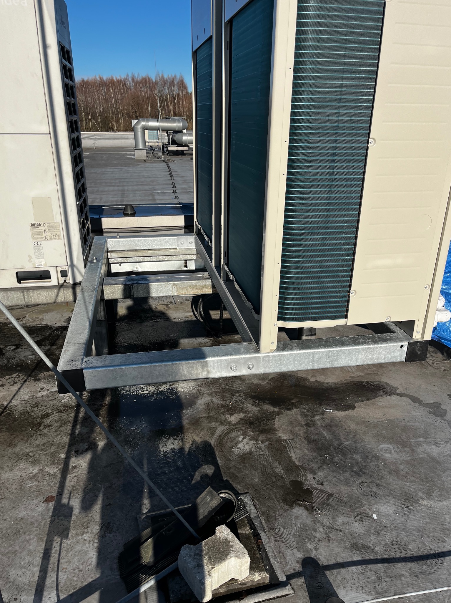 Montaż klimatyzacji na dachu budynku. Widoczna stalowa konstrukcja wsporcza pod agregat, elementy instalacji i fragment dachu. Słoneczny dzień.