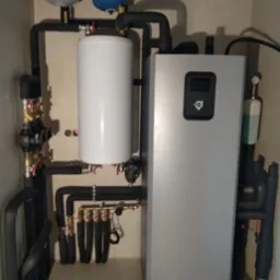 Zainstalowana szara jednostka z wyświetlaczem i białym zbiornikiem, z widocznymi rurami i osprzętem w kotłowni.