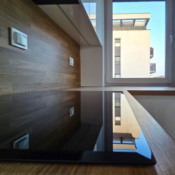 PRONEO SPÓŁKA Z OGRANICZONĄ ODPOWIEDZIALNOŚCIĄ - Nowoczesna kuchnia z drewnianym blatem i płytą indukcyjną, odbijająca w oknie widok na budynek mieszkalny w słoneczny dzień. Minimalistyczny design.