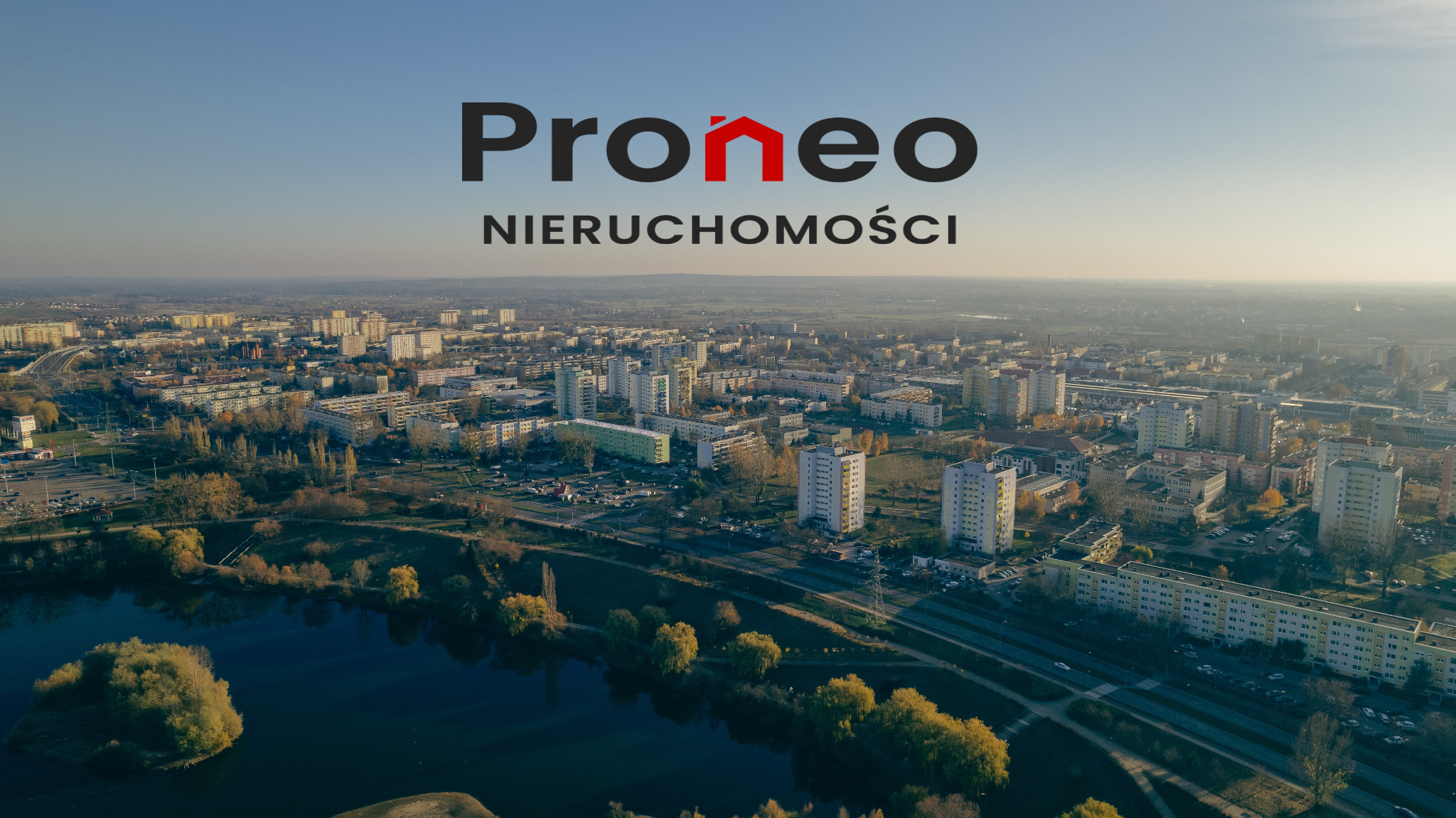 Panorama Konina z lotu ptaka, z widocznym jeziorem i blokami mieszkalnymi, logo Proneo Nieruchomości na pierwszym planie.