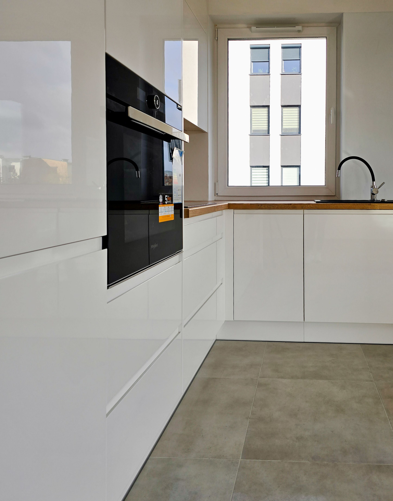 Nowoczesna, biała kuchnia z zabudową, piekarnikiem i widokiem na budynek z okna. Minimalistyczny design, szare płytki podłogowe, drewniany blat.