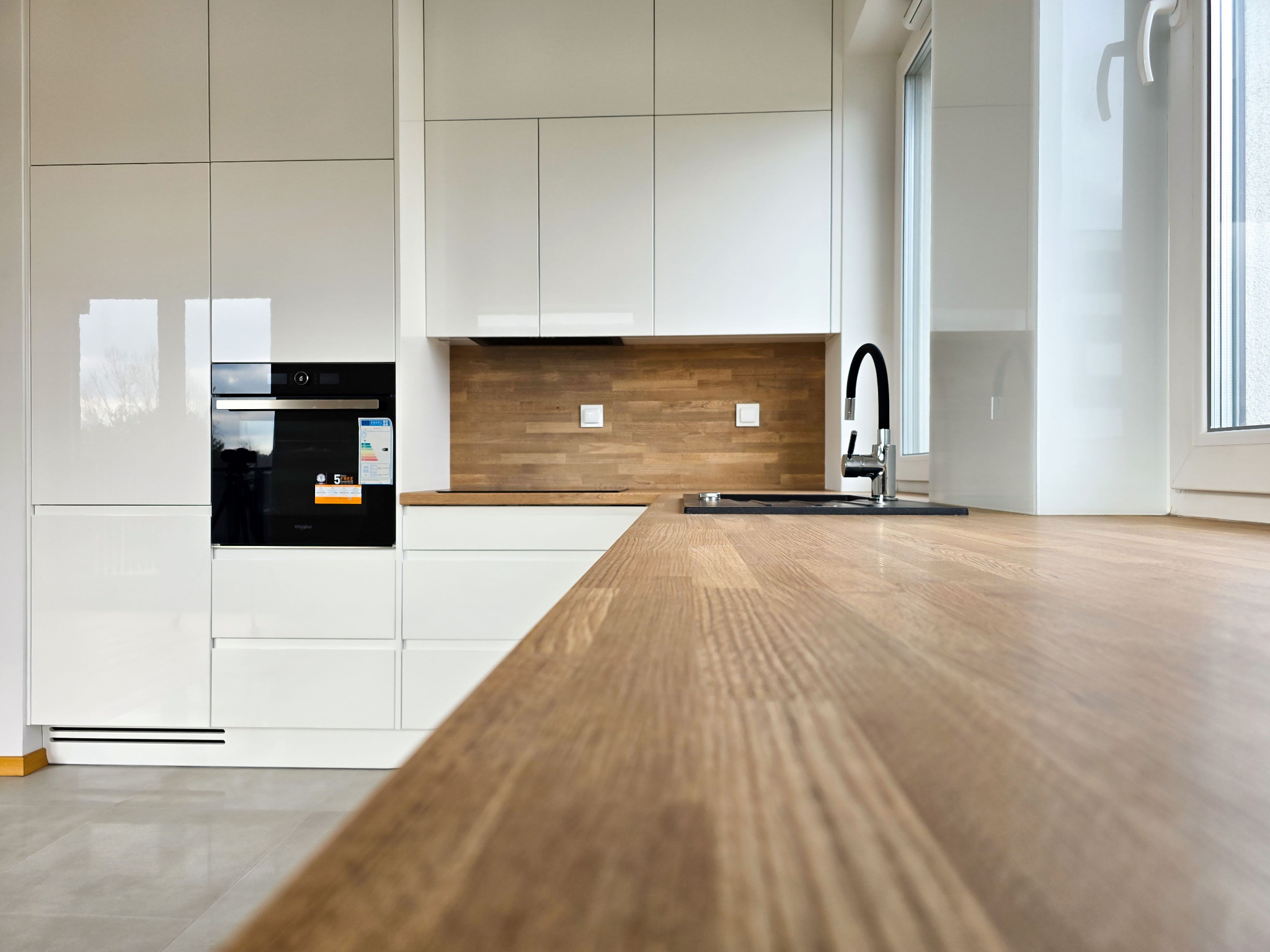 Nowoczesna, jasna kuchnia z białymi, lakierowanymi frontami, drewnianym blatem i panelem ściennym. Widoczny piekarnik i bateria kuchenna. Minimalistyczny design.