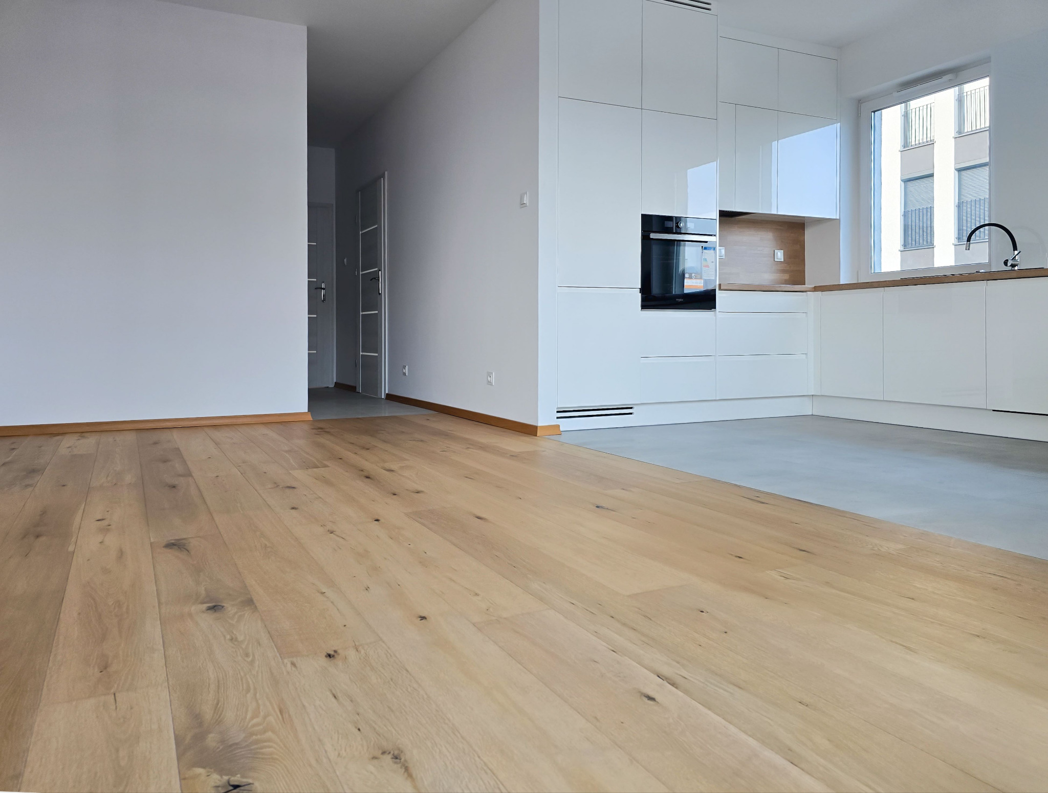 Jasne, drewniane panele podłogowe w nowym mieszkaniu z białą zabudową kuchenną i widokiem na budynek zza okna. Minimalistyczne wnętrze.
