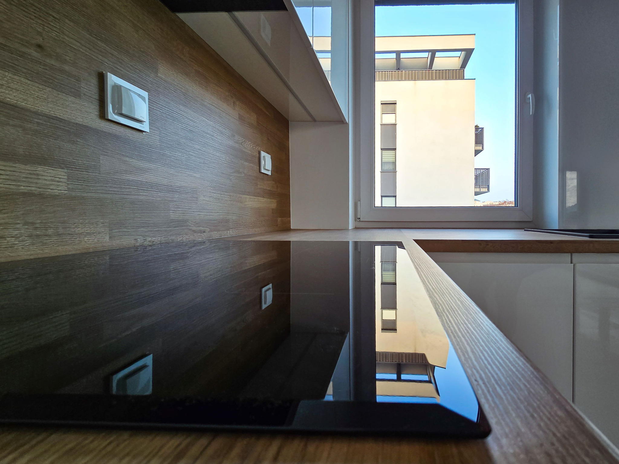 Nowoczesna kuchnia z drewnianym blatem i płytą indukcyjną, odbijająca w oknie widok na budynek mieszkalny w słoneczny dzień. Minimalistyczny design.