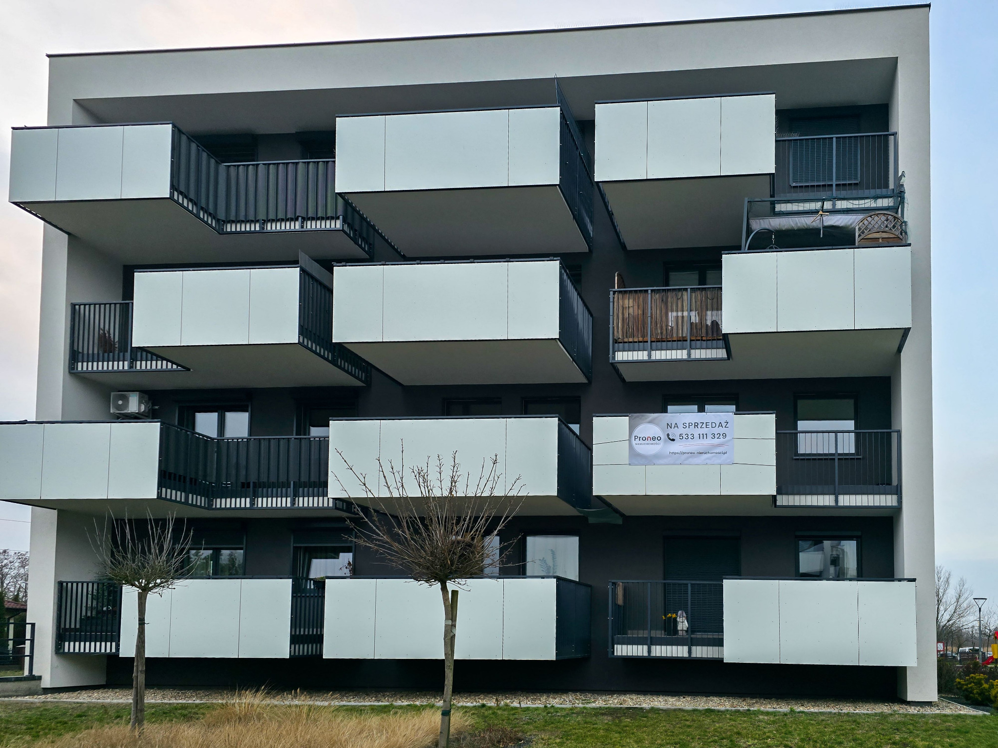 Nowoczesny apartamentowiec z balkonami w kształcie kostek. Na jednym z balkonów widoczna tabliczka 'Na sprzedaż' z numerem telefonu. Minimalistyczna architektura.