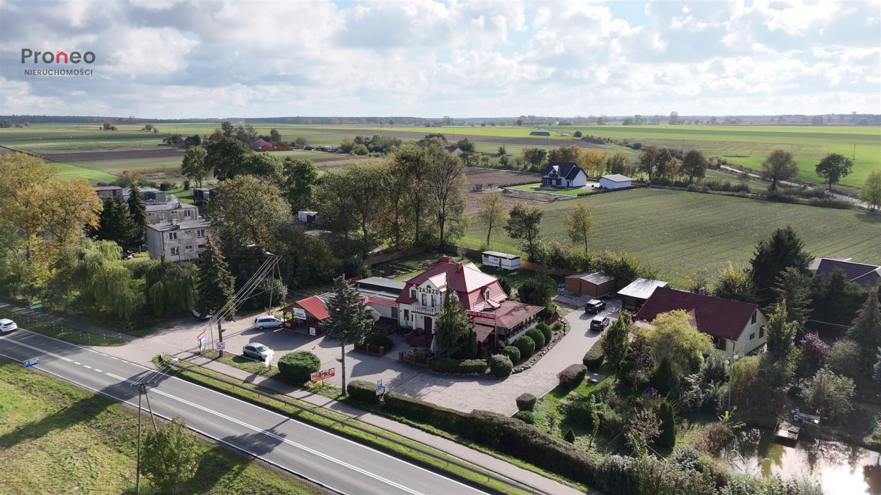 Panoramiczny widok z lotu ptaka na wiejski krajobraz z budynkami mieszkalnymi, polem uprawnym i drogą. W tle widoczne chmury i logo agencji nieruchomości.