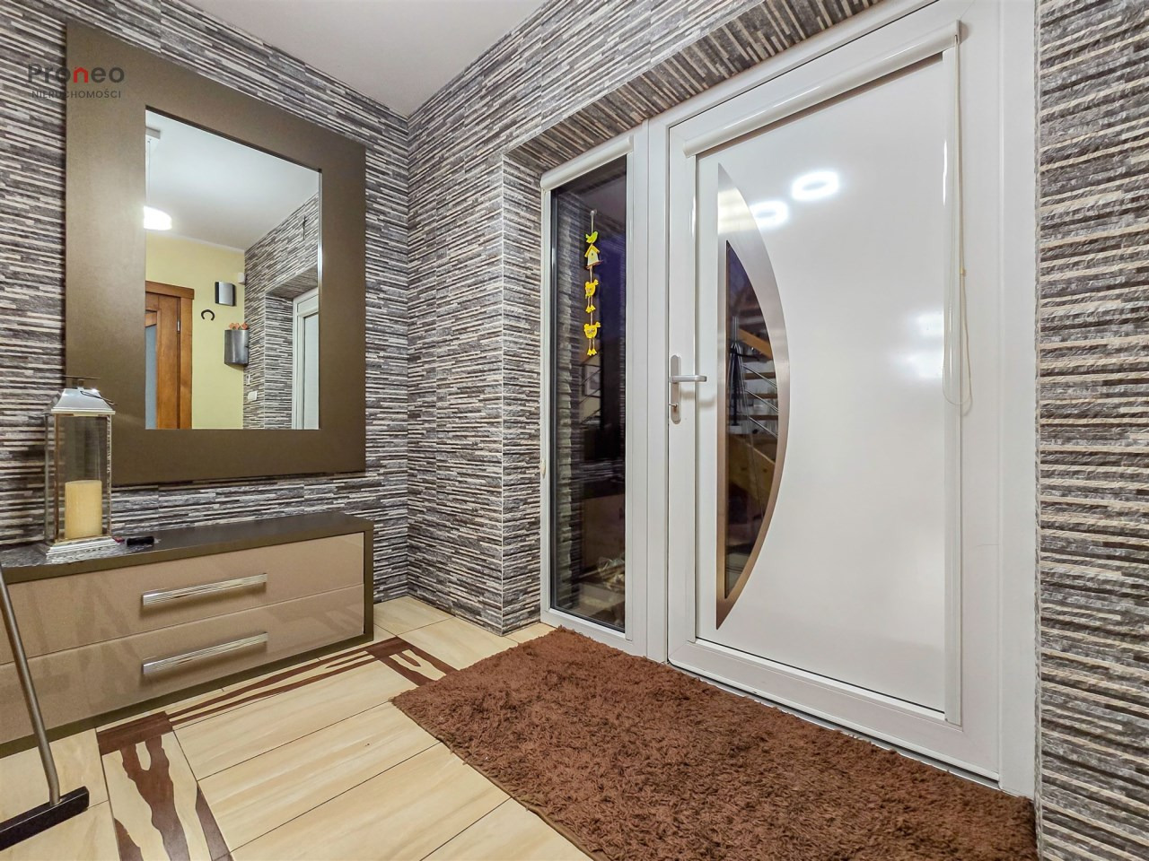 Nowoczesny przedpokój z lustrem, komodą i białymi drzwiami wejściowymi. Dekoracyjna ściana z kamienia, brązowy dywanik. Widok na wnętrze mieszkania.