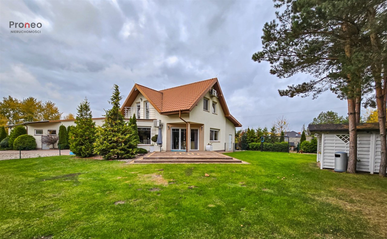 Jasny dom jednorodzinny z czerwoną dachówką, balkonem i dużym ogrodem. Widok z trawnika w pochmurny dzień. W tle budynek gospodarczy i drzewa.