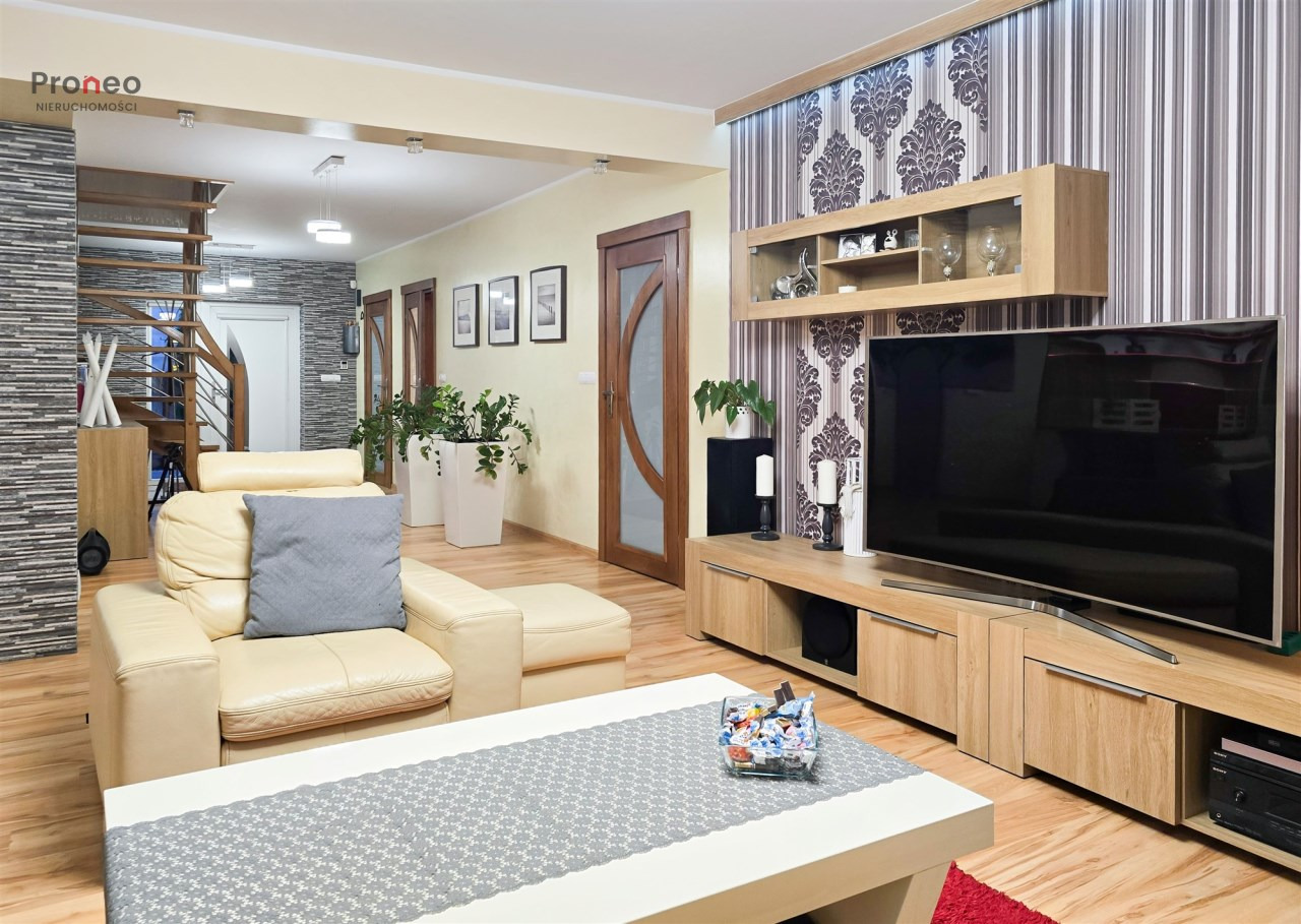 Jasny salon z beżową sofą, drewnianymi schodami i telewizorem na tle wzorzystej tapety. Na stole dekoracyjny bieżnik i szklane naczynie ze słodyczami.