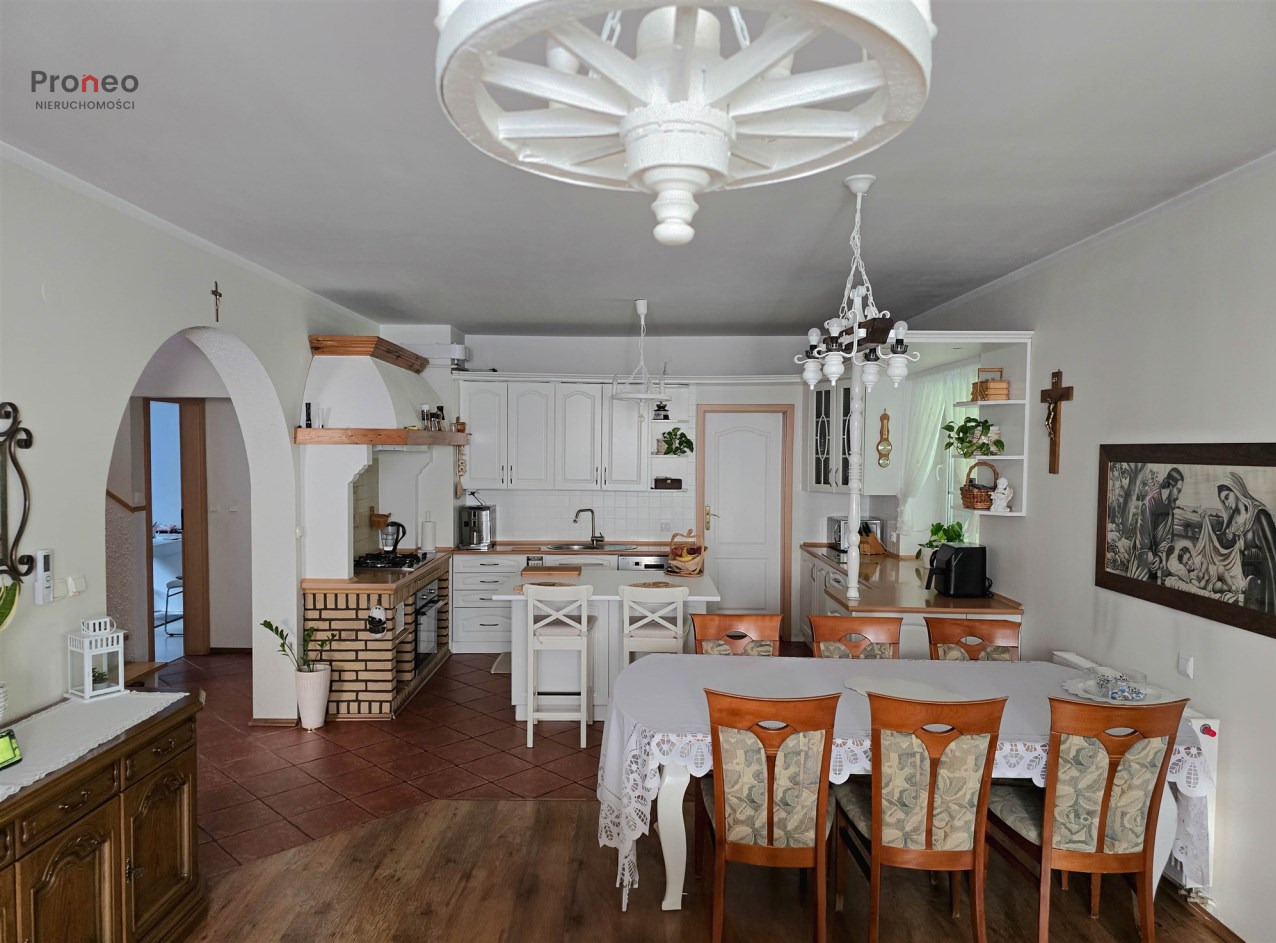 Jasne wnętrze: kuchnia z ceglaną zabudową, białe meble, drewniany stół z krzesłami. Nietypowa lampa sufitowa w kształcie koła od wozu. Widok na salon.