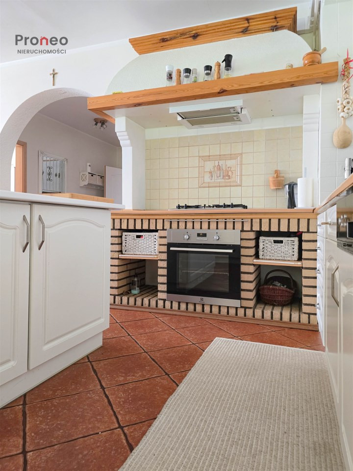 Rustykalna kuchnia z ceglaną zabudową piekarnika, białymi szafkami i terakotą na podłodze. W tle łukowe przejście do innego pomieszczenia. Widoczny dywanik.