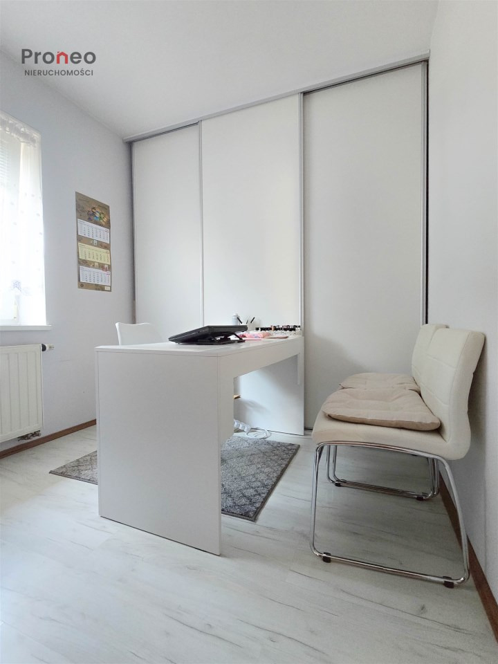 Jasne biuro z białym biurkiem, krzesłem i szafą przesuwną. Minimalistyczny wystrój, dywanik na podłodze. Widok na wnętrze biura nieruchomości.